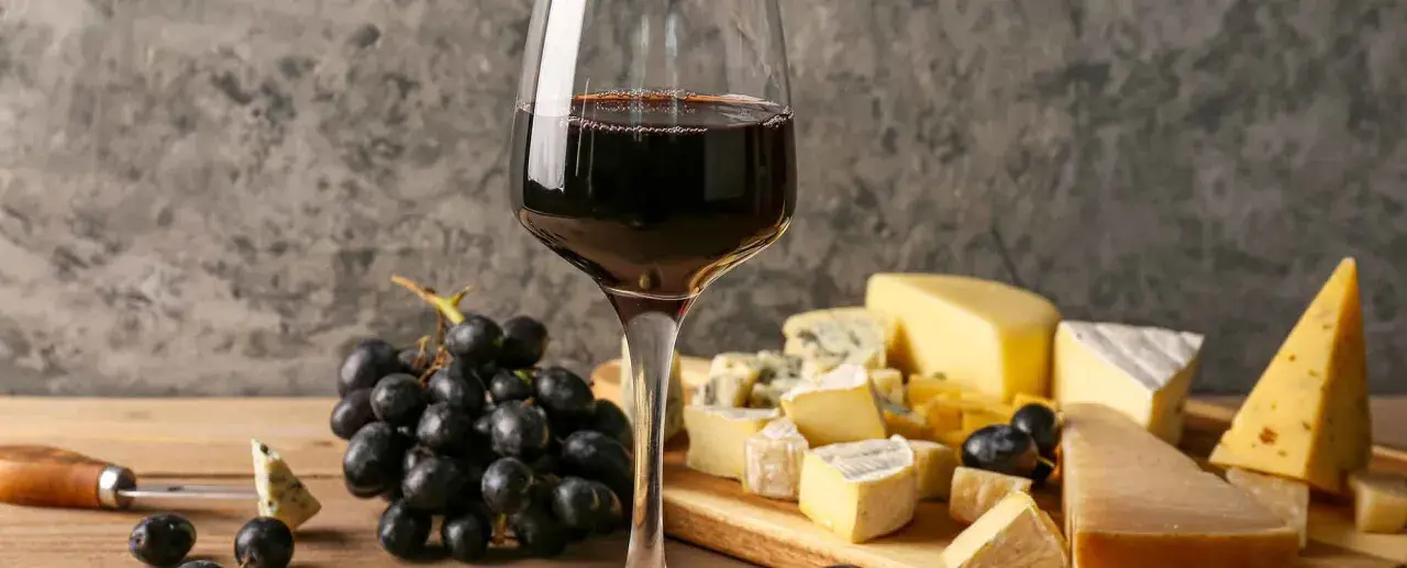 Un bicchiere di vino rosso accompagna un invitante tagliere di formaggi misti, con uva nera e un coltello da formaggio.