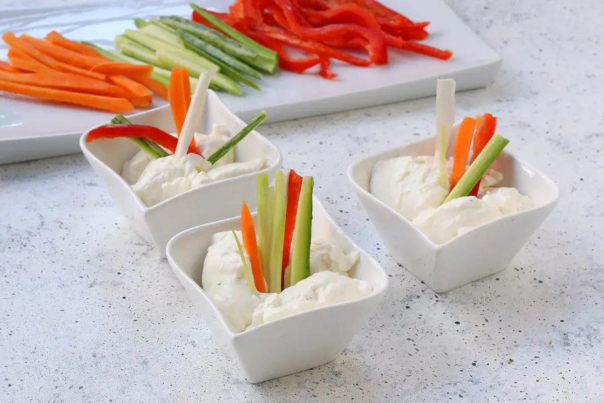 Tre ciotoline bianche con mousse di caprino, guarnite con bastoncini di verdure fresche. Sullo sfondo, verdure tagliate a julienne.