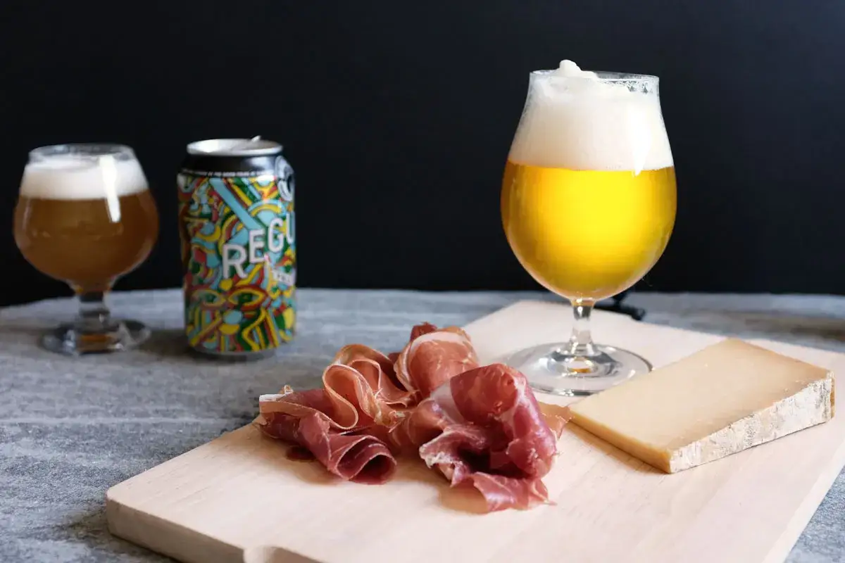 Un bicchiere di birra dorata, una lattina colorata e un tagliere con prosciutto e formaggio. Un abbinamento perfetto di birra e formaggio.