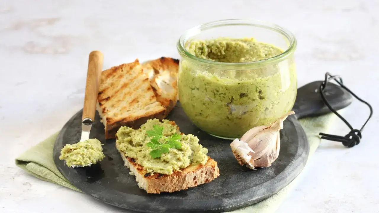 Un vasetto di crema spalmabile verde, pane tostato, aglio e un cucchiaio con la crema. Perfetta per tartine, questa crema è un'ottima alternativa alla panna da cucina.