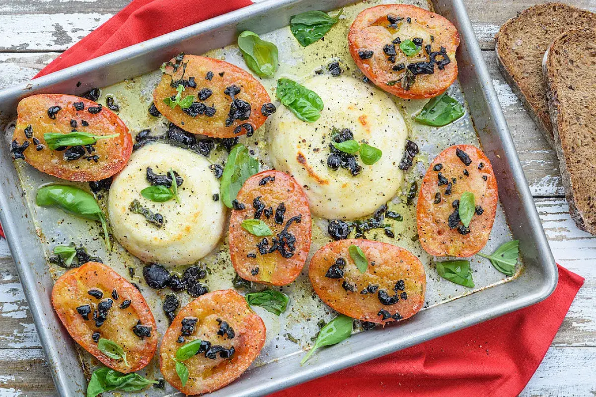 Primo sale al forno con pomodori, olive e basilico su teglia metallica, accompagnato da fette di pane integrale.