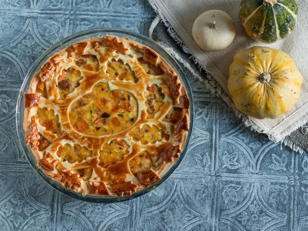 Torta salata zucca salsiccia e stracchino, dorata e invitante, con un motivo a foglie sulla crosta. Accanto, zucche decorative.
