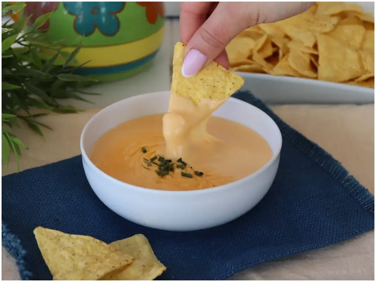 Una mano intinge un triangolo di mais nella cremosa salsa cheddar, guarnita con erba cipollina.