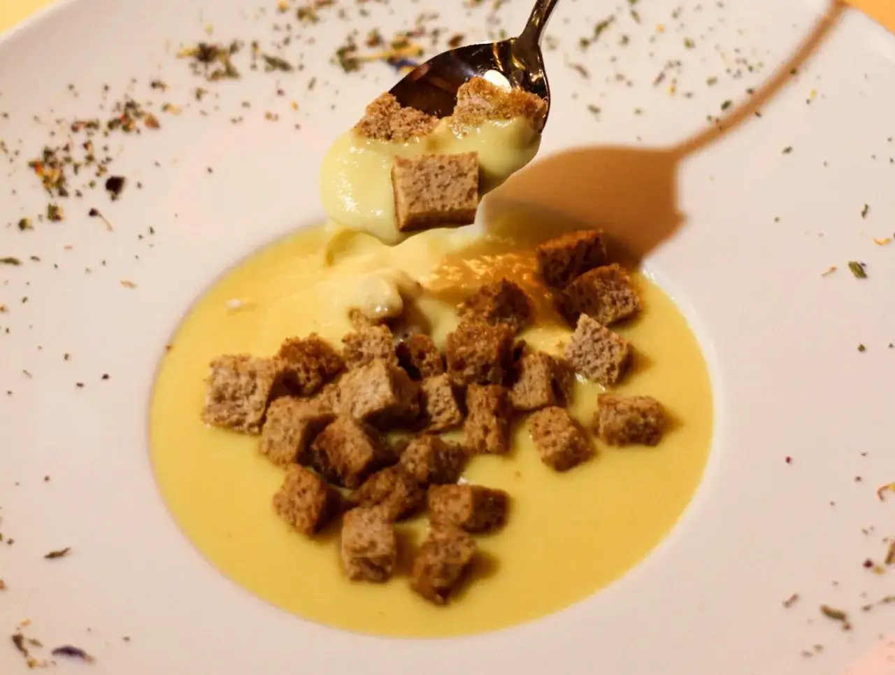 Una cucchiaiata di cremosa fonduta di formaggio con crostini dorati, servita in un piatto bianco decorato con erbe aromatiche.