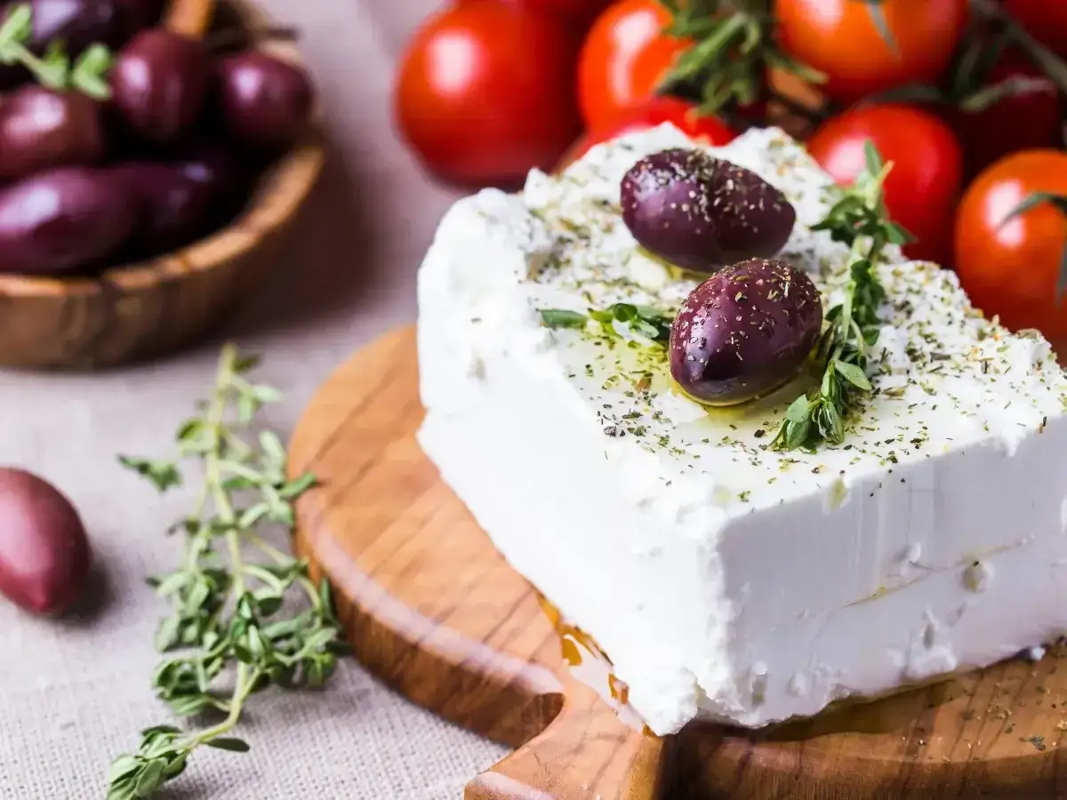 Un blocco di la feta condito con erbe e olive, pronto per una fresca insalata greca con pomodori sullo sfondo.
