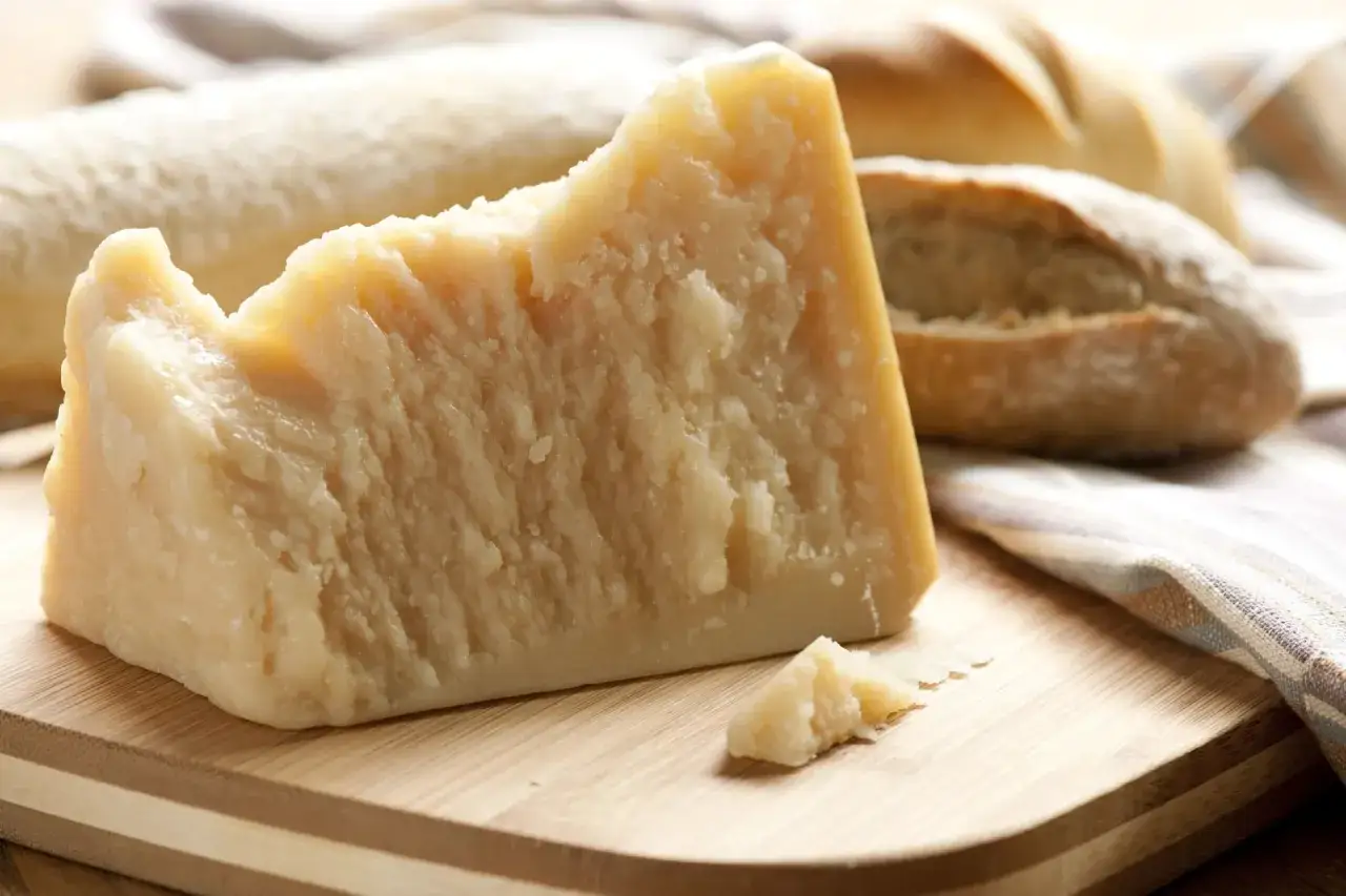 Un pezzo di parmigiano stagionato su un tagliere di legno, con pane sullo sfondo. Un alimento gustoso, forse un aiuto contro l'artrosi.